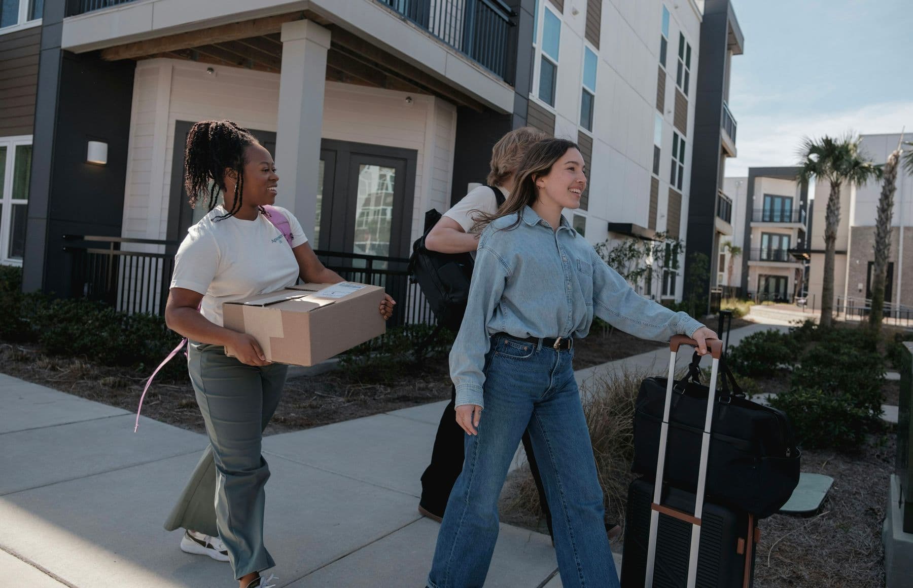 Three friends carrying moving boxes and a suitcase into their new apartment building